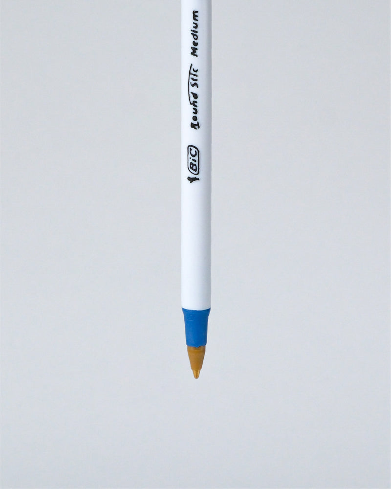 A BIC Liquid Stick Medium pen shown vertically against a light gray background, featuring a white barrel with black printed branding and a blue grip accent near the tip.