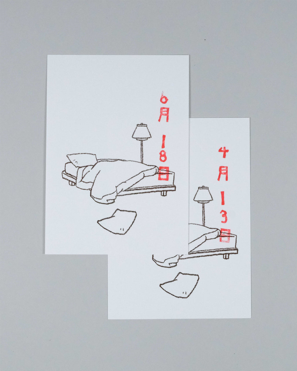 Two white art prints with minimalist line drawings of a bed, lamp, and clothing items, annotated with red Japanese dates, displayed on a neutral gray background.
