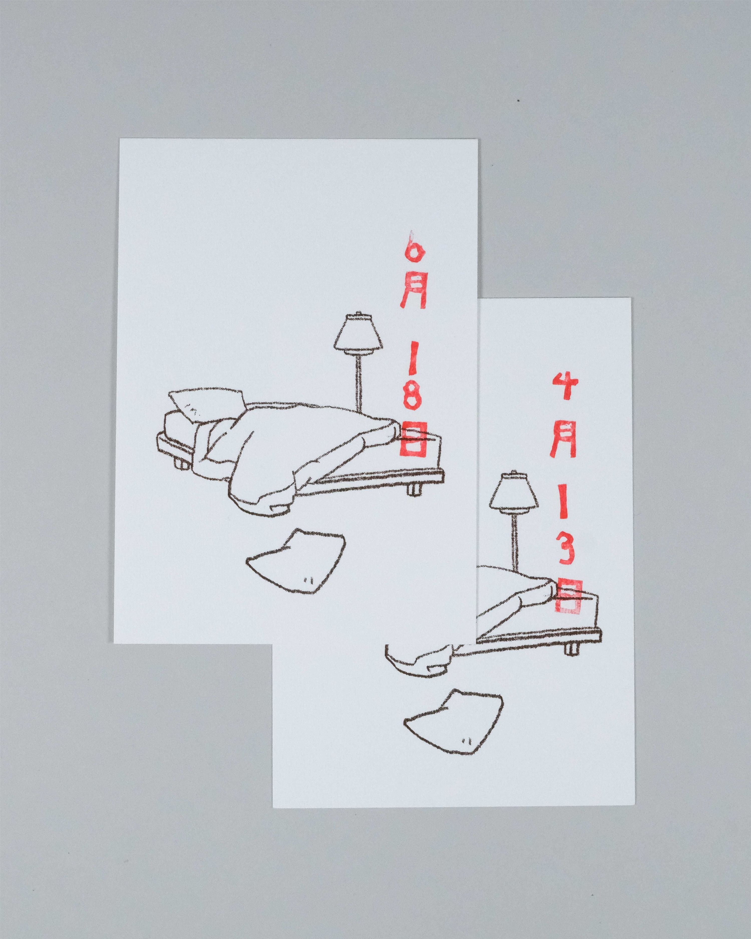 Two white art prints with minimalist line drawings of a bed, lamp, and clothing items, annotated with red Japanese dates, displayed on a neutral gray background.