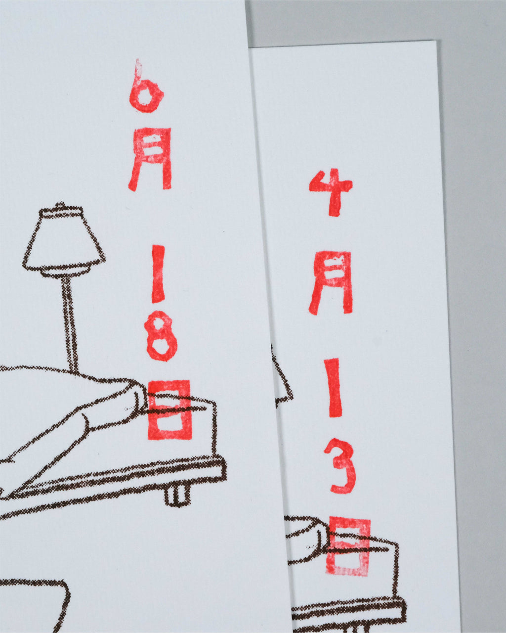 Two white rectangular cards or prints with minimalist line drawings and red stamped Japanese characters and dates, shown overlapping on a light gray background.