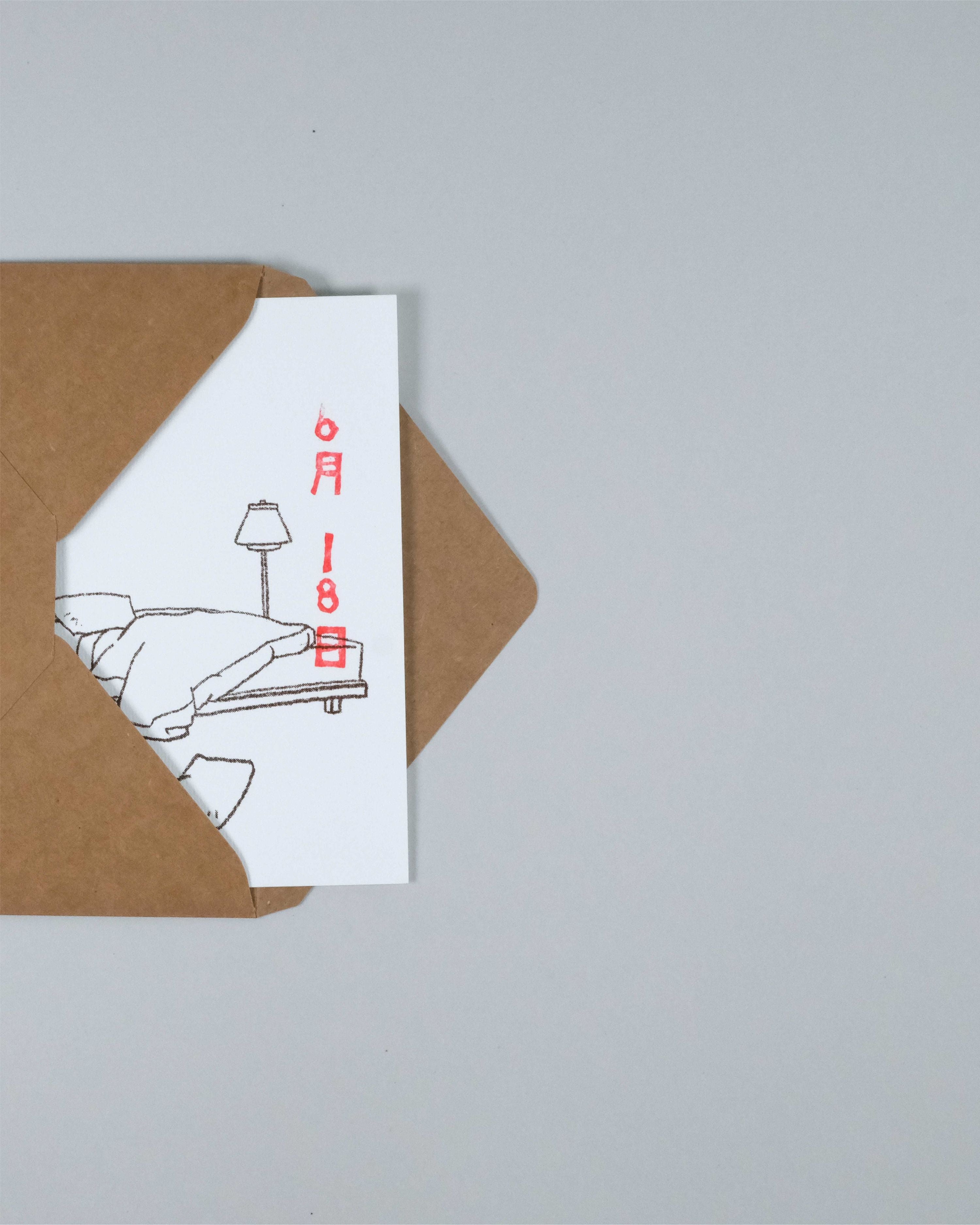 A white greeting card with a minimalist black line drawing of a person turning off a lamp, with red text reading '6月18日', partially tucked inside a brown kraft paper envelope on a light gray background.