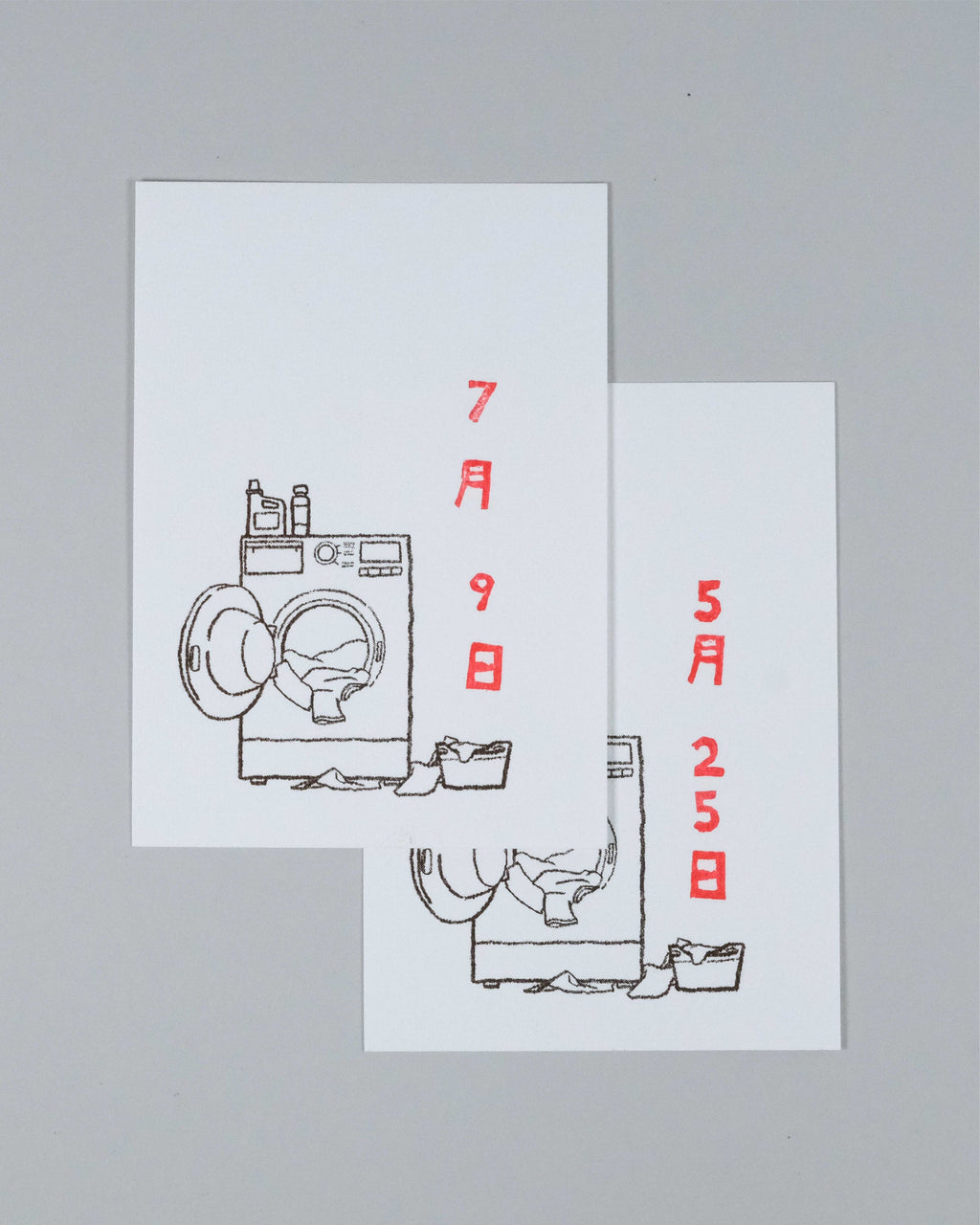 Two overlapping white greeting cards featuring minimalist black line drawings of washing machines and laundry baskets, with red Japanese text displaying dates, set against a solid gray background.