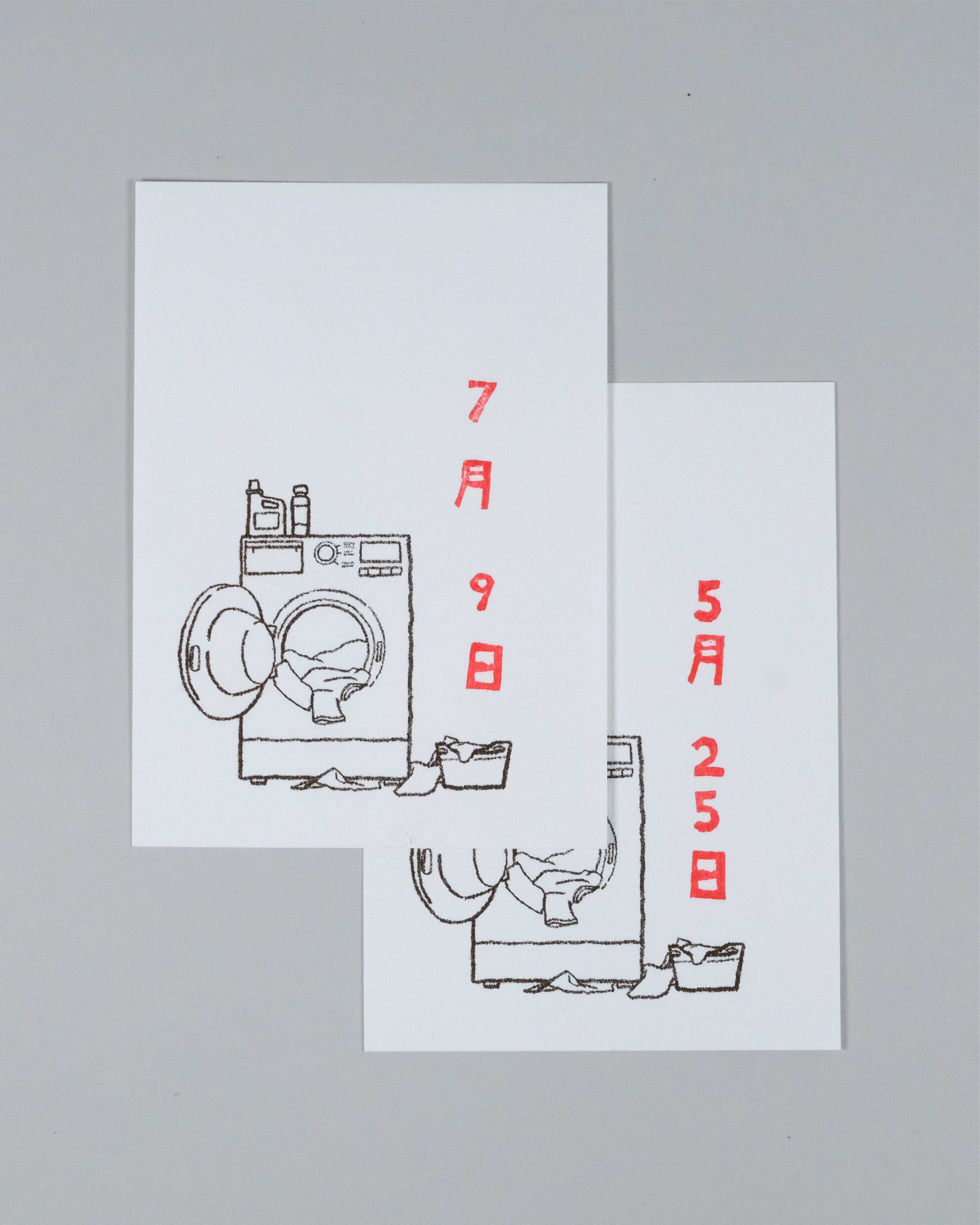 Two overlapping white greeting cards featuring minimalist black line drawings of washing machines and laundry baskets, with red Japanese text displaying dates, set against a solid gray background.