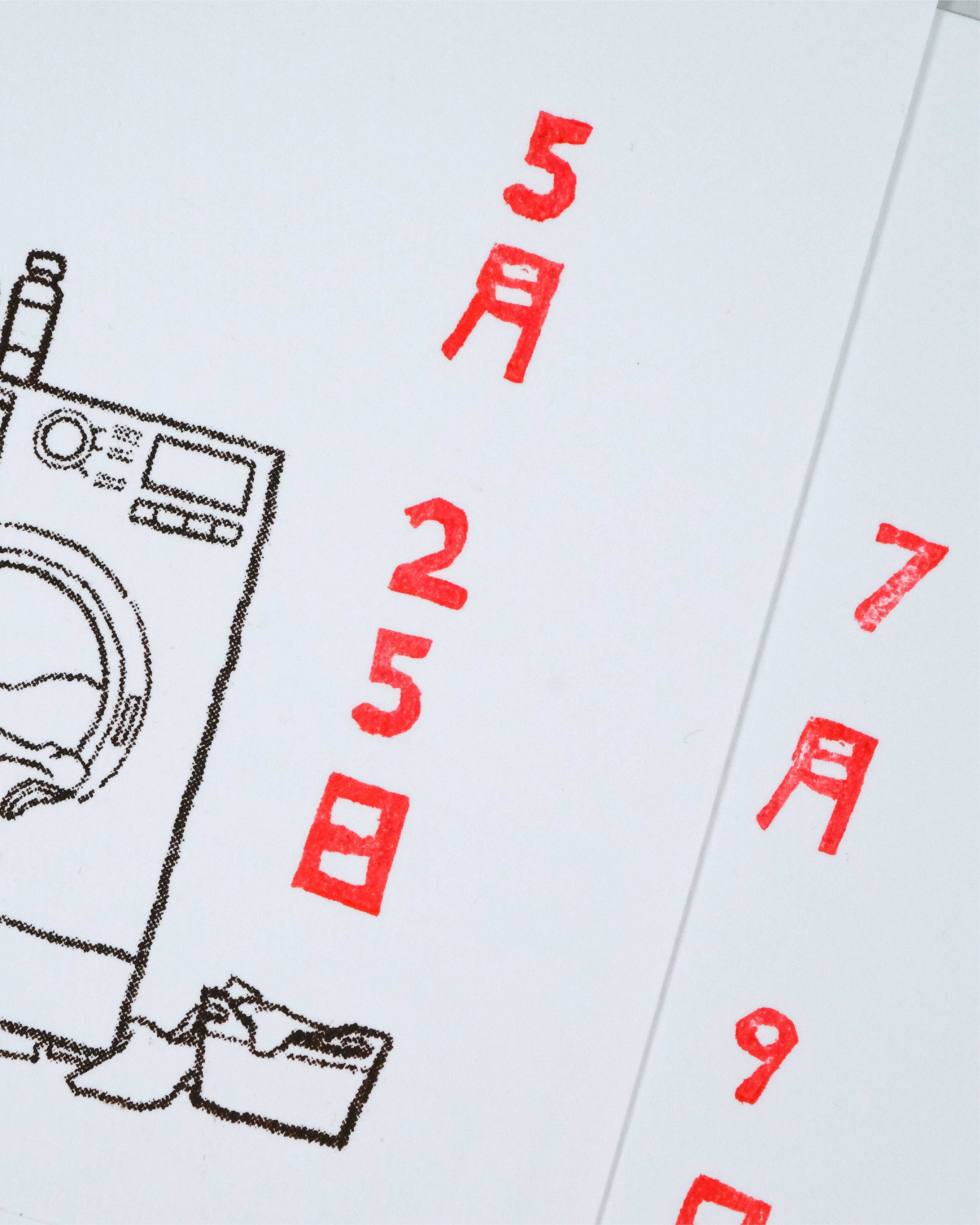 A close-up of a white paper calendar or planner page with a black line drawing of a washing machine and clothes basket, alongside red Japanese numerals showing dates 5月25日 and 7月9日.