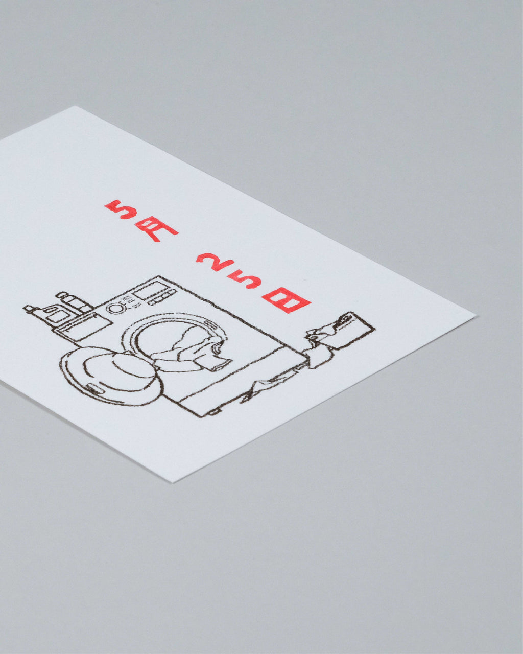 A white rectangular card or label with a black line drawing of a washing machine and folded clothes, plus red Japanese text, photographed on a light gray background.