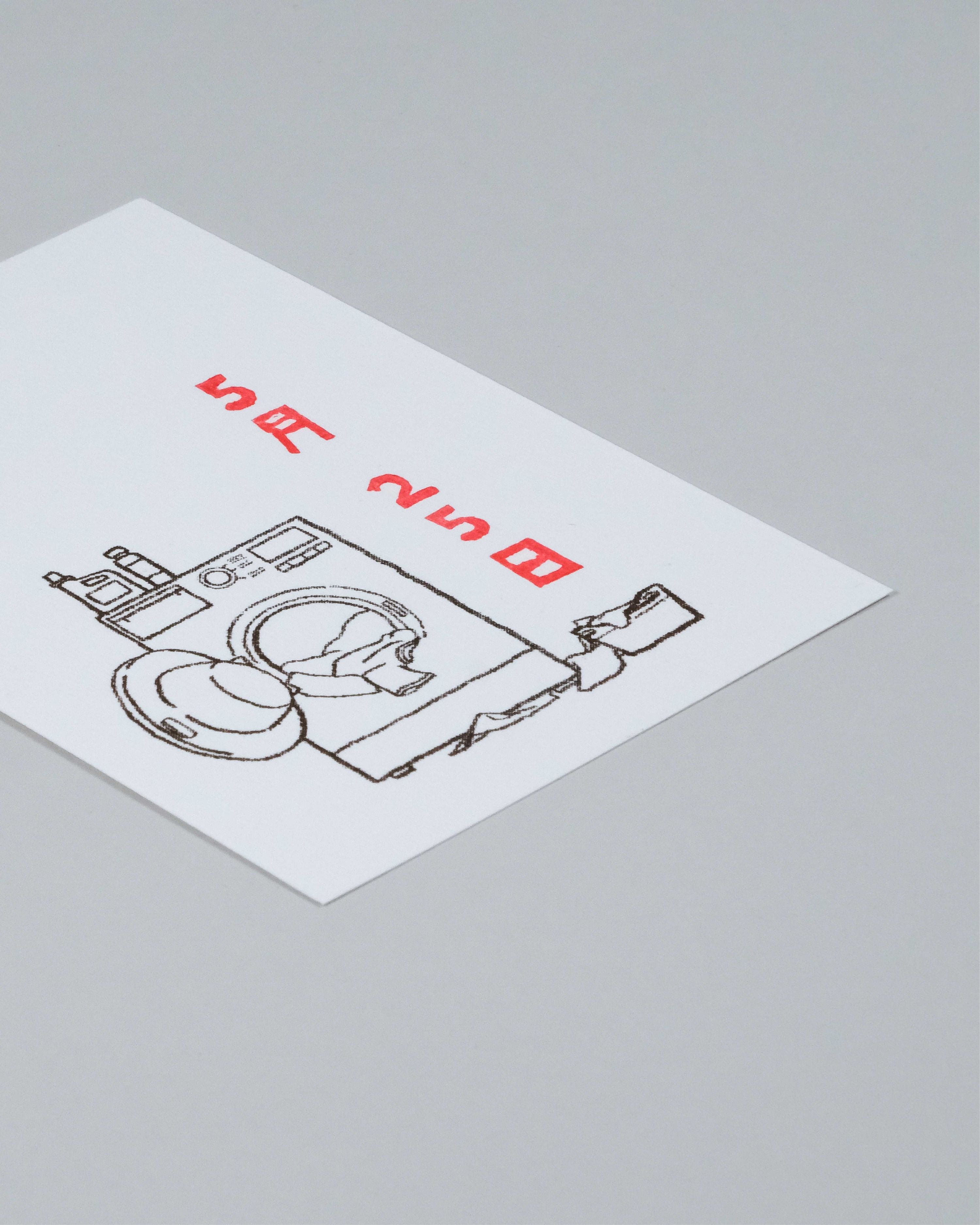 A white rectangular card or label with a black line drawing of a washing machine and folded clothes, plus red Japanese text, photographed on a light gray background.