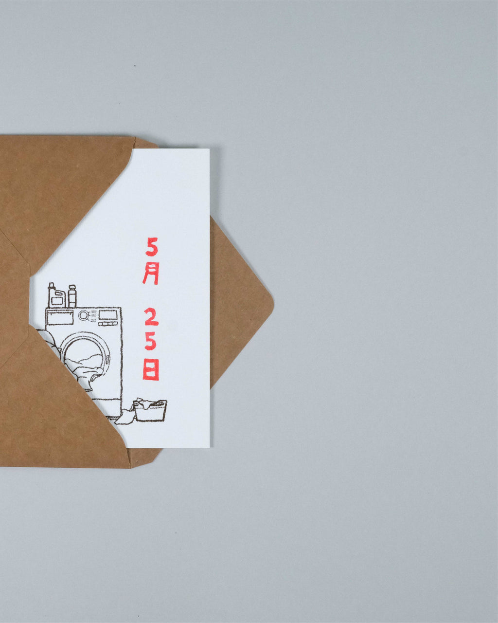 A white greeting card with a minimalist black line drawing of a washing machine and red vertical Japanese characters, partially tucked into a brown kraft envelope on a light gray background.