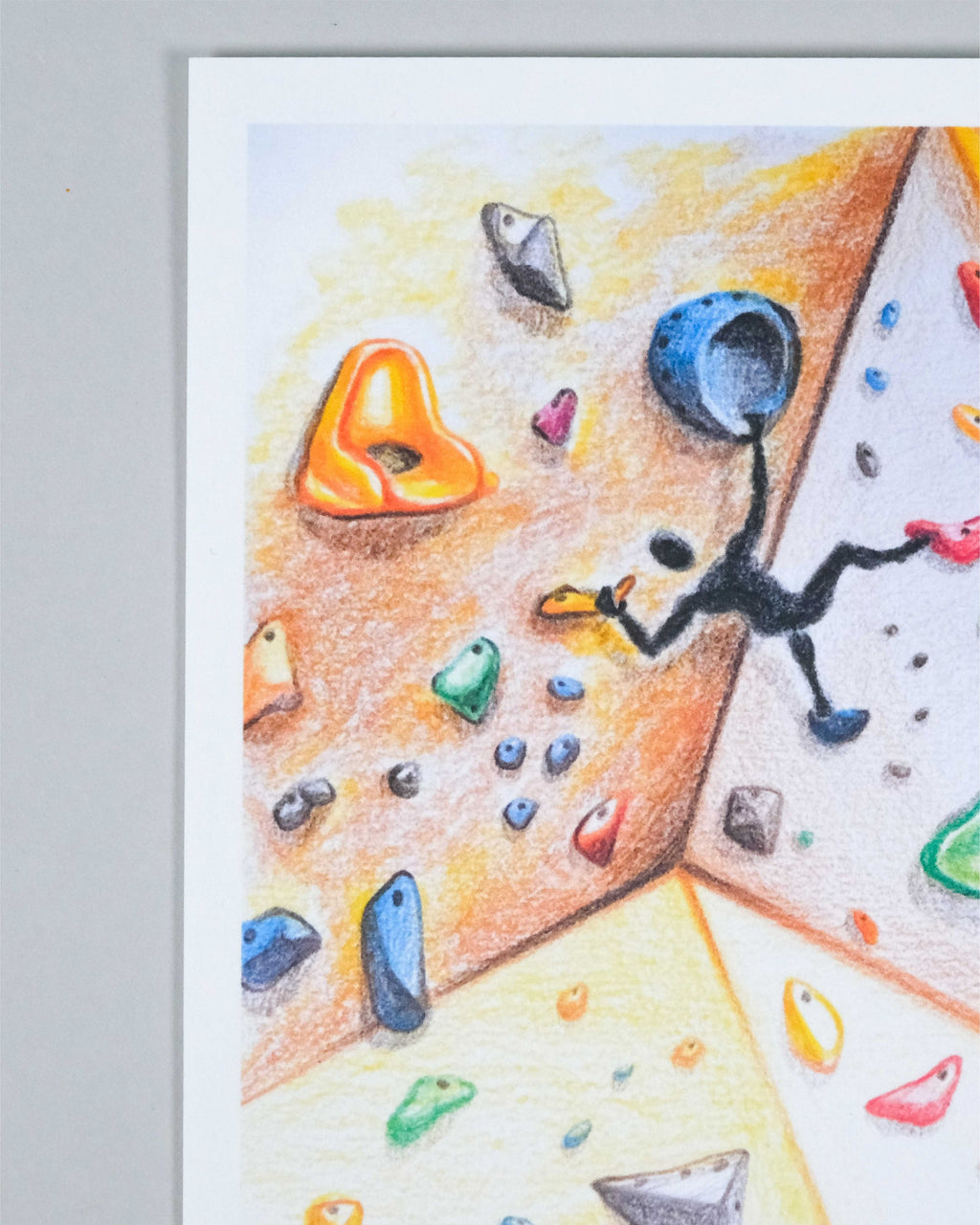 A close-up of a colorful climbing wall drawing or print, showing climbing holds in orange, blue, green, red, and gray on a textured background, with a black stick-figure climber. The print is photographed against a light gray background.
