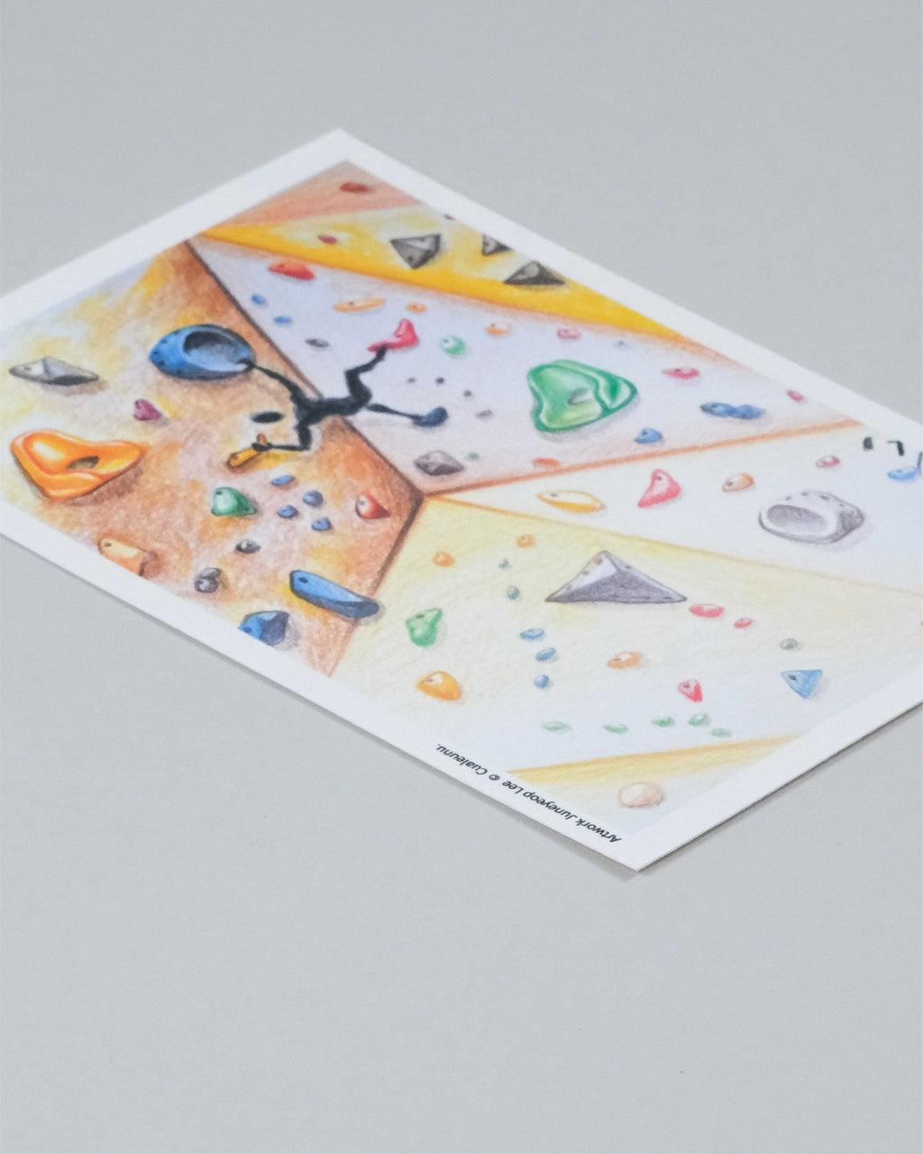A rectangular art print featuring a colorful climbing wall illustration, photographed at an angle on a neutral gray background.