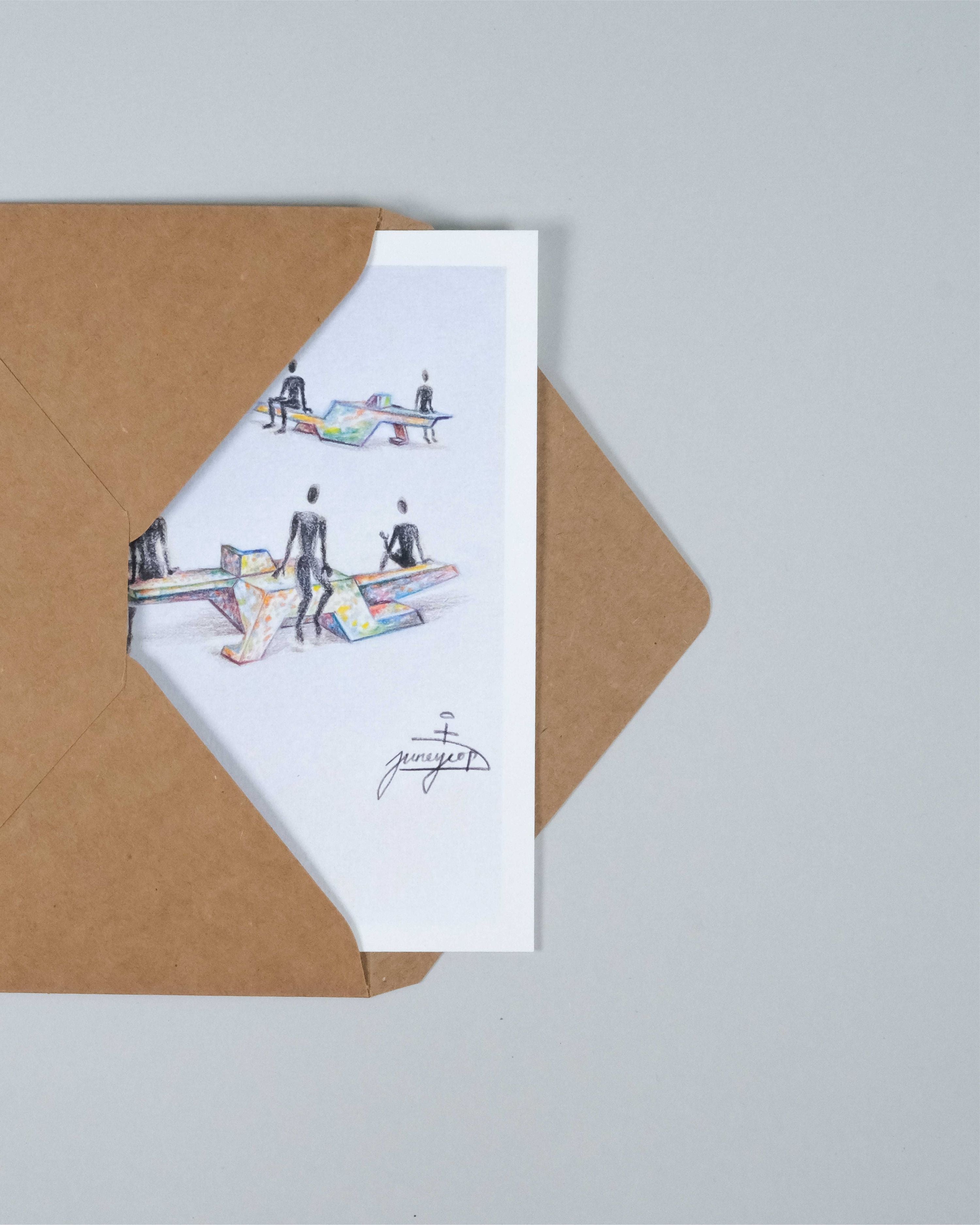 A white art print card featuring a colored pencil sketch of people at a table, partially tucked inside a brown kraft paper envelope on a light gray background.