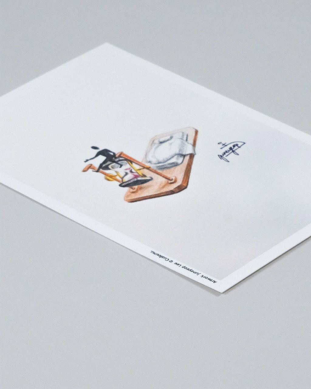 A minimalist art print on white paper, featuring a small illustration of a figure with a bicycle and a bicycle frame, photographed on a neutral gray background.