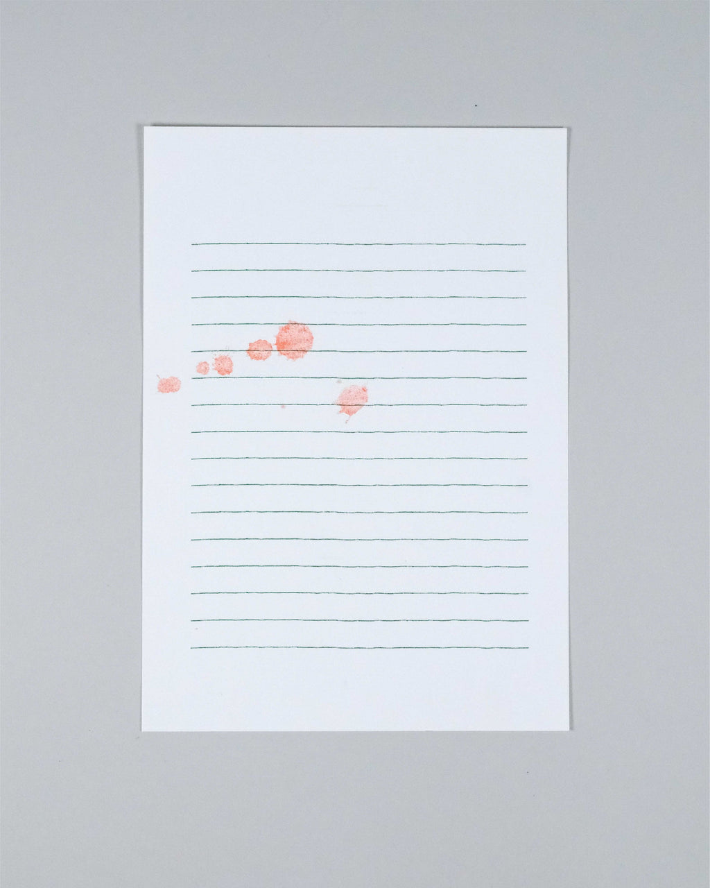 A vertical white lined notepad with faint green horizontal lines and a few scattered coral-pink watercolor dots, photographed against a light gray background.