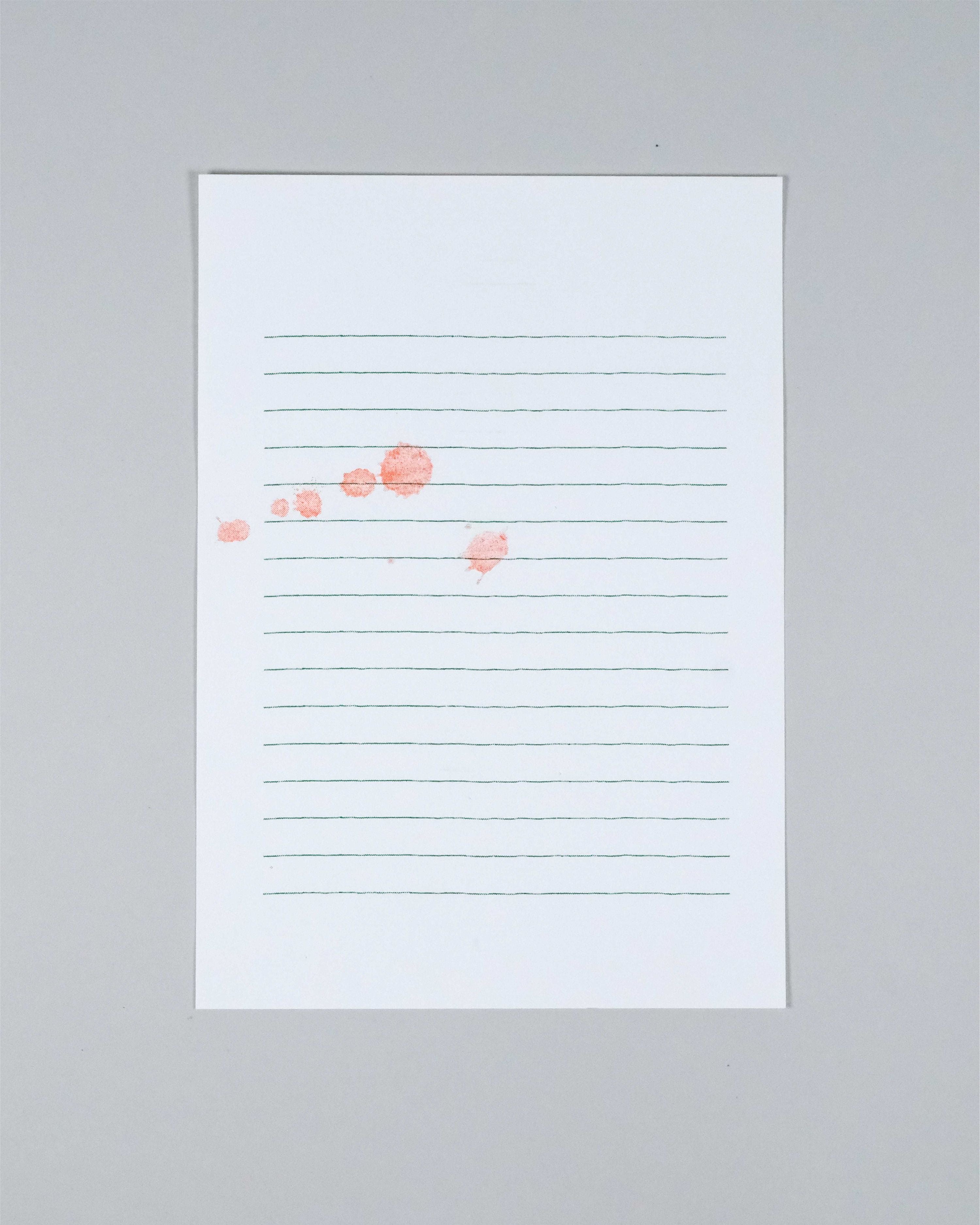 A vertical white lined notepad with faint green horizontal lines and a few scattered coral-pink watercolor dots, photographed against a light gray background.