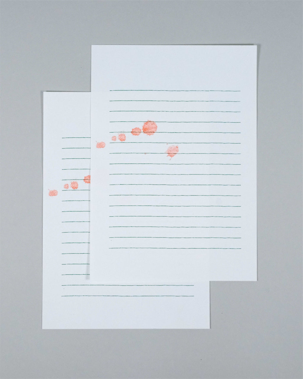 Two sheets of lined white paper with horizontal blue lines and scattered coral-pink circular marks, arranged on a plain light gray background.