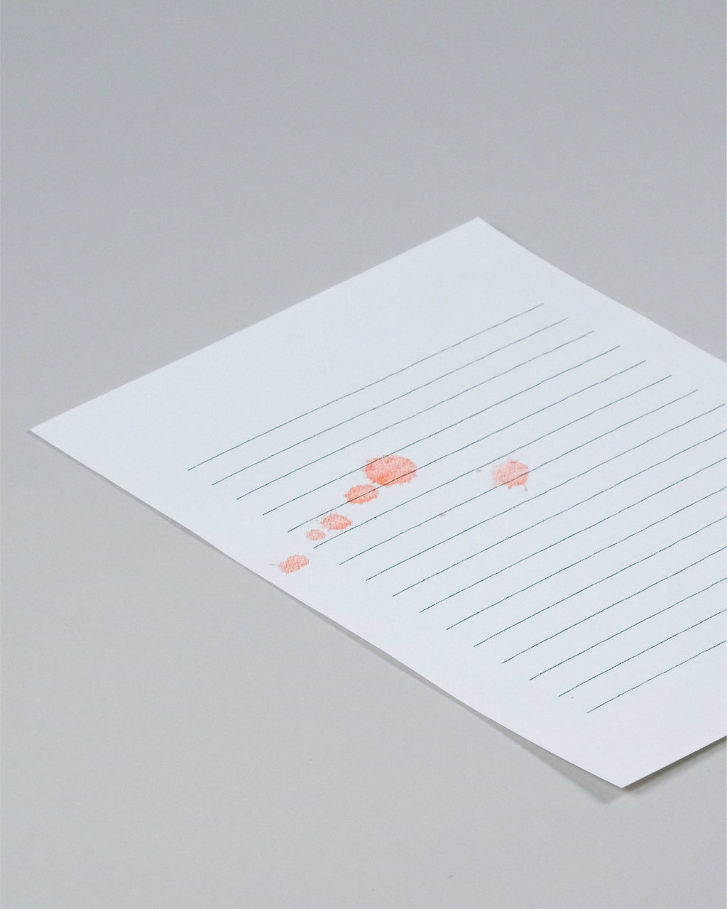 A square white paper card with horizontal gray lines and a small coral-pink abstract graphic near the lower-left area, photographed on a light gray background.