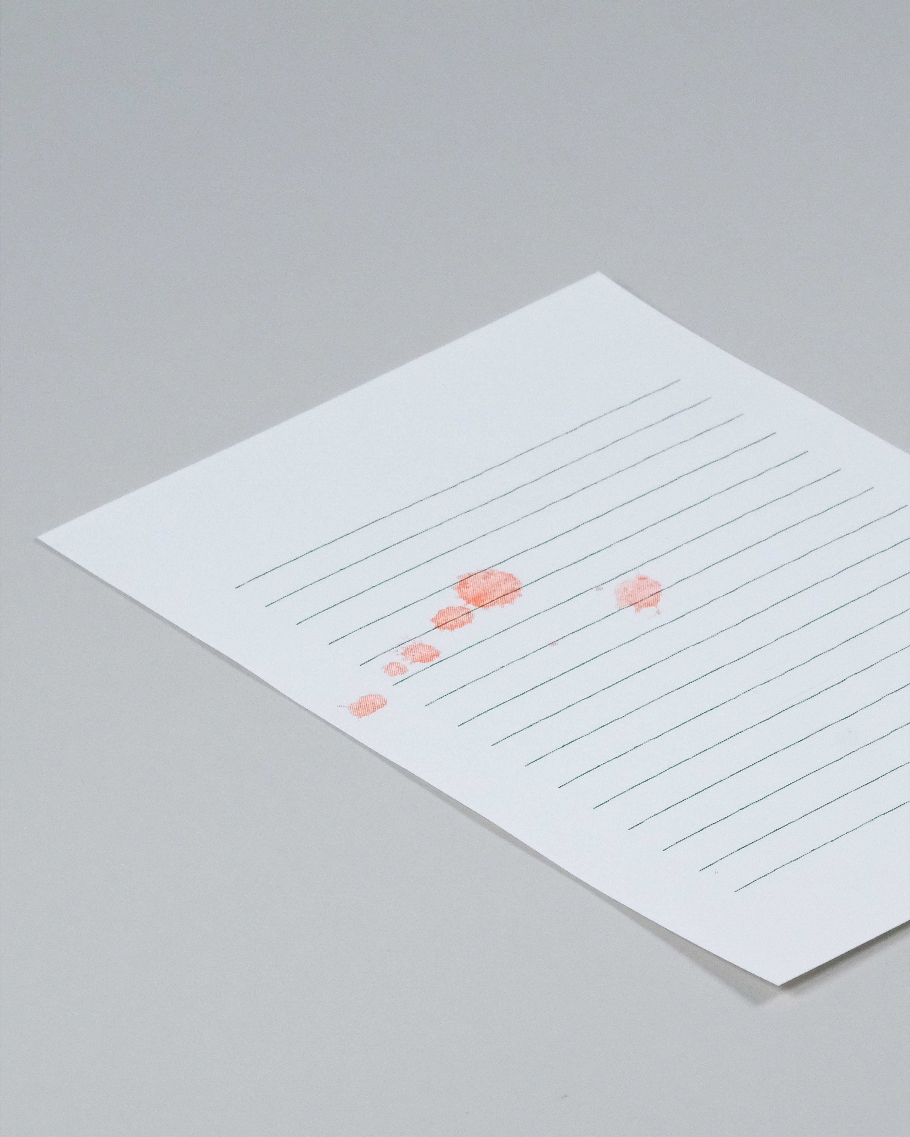 A square white paper card with horizontal gray lines and a small coral-pink abstract graphic near the lower-left area, photographed on a light gray background.