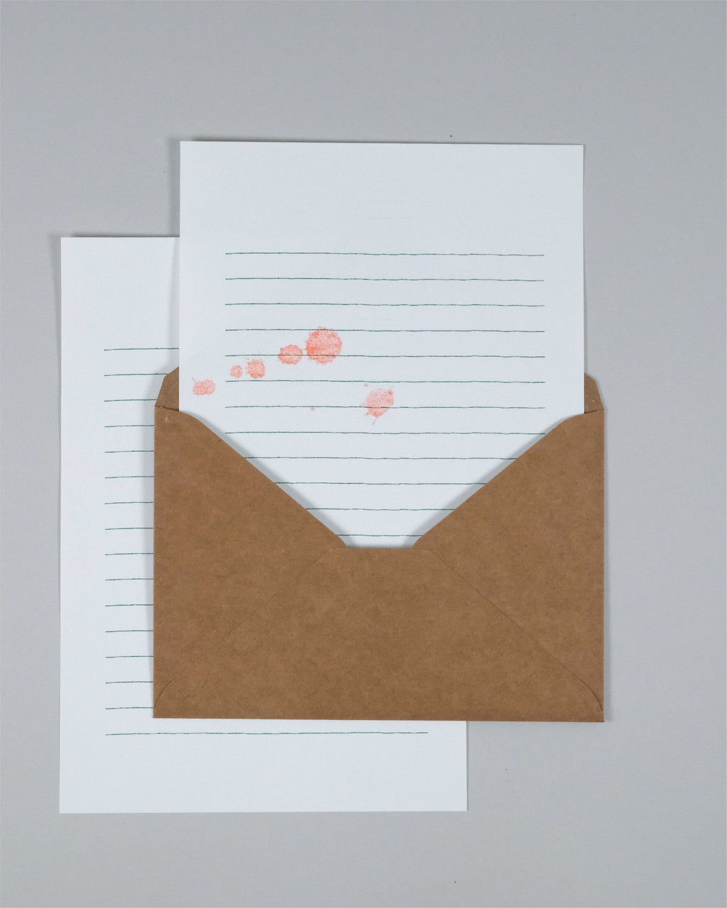 A kraft brown envelope with a white lined card partially pulled out, decorated with small pink heart-shaped stamps, set against a neutral gray background.