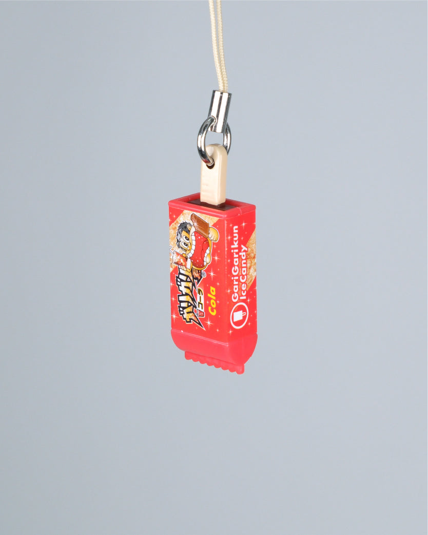 A red snack-shaped keychain or charm with printed graphics and a cream cord attached by a silver metal ring, shown against a plain light gray background.