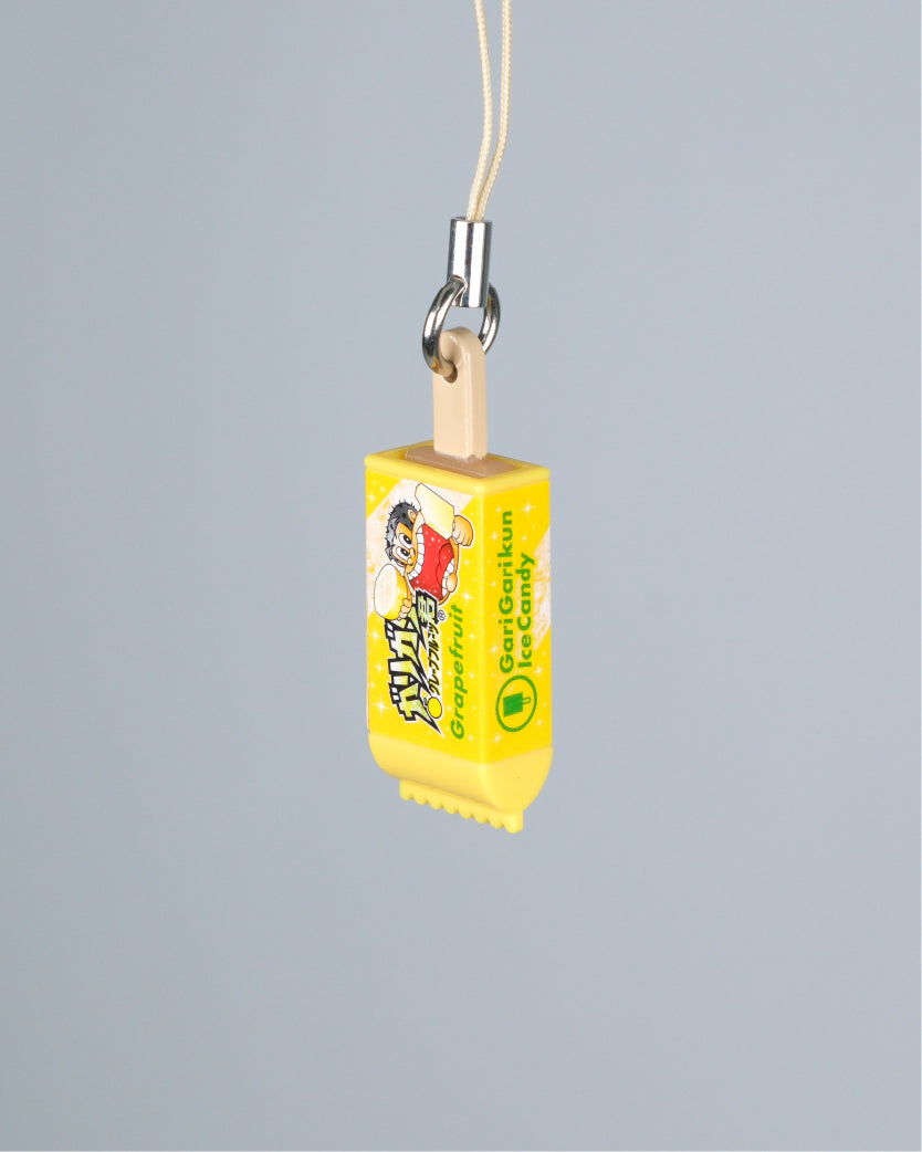 A miniature yellow toy candy bar charm with a wooden stick, metal clasp, and a thin beige hanging cord, shown against a solid gray background.