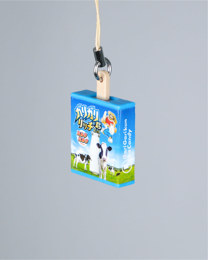 A small rectangular blue product box with cartoon cow illustrations and Chinese text, hanging from a beige cord and black metal clip against a plain light gray background.