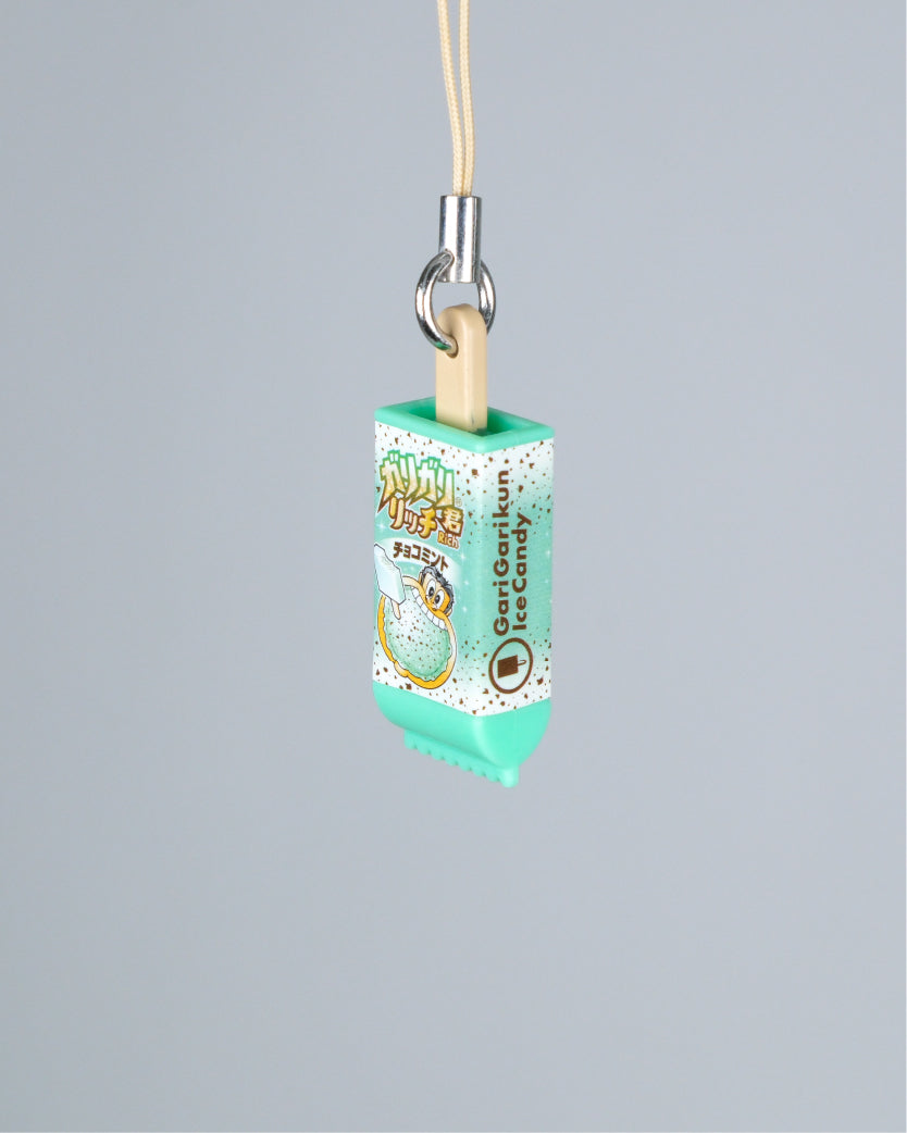 A mint green and white ice cream-shaped phone strap charm with Japanese text and a cartoon illustration, suspended by a beige cord with a silver metal clasp against a solid gray background.