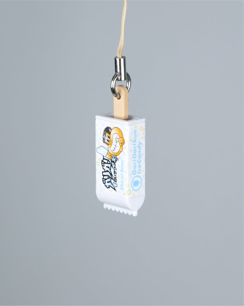 A miniature white candy wrapper charm with a cartoon character and light blue text, attached to a beige string and silver metal clasp, shown against a solid gray background.