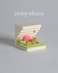 Small model of a building with a cherry blossom tree and picnic setup on a gray background, labeled 'spring edition'.