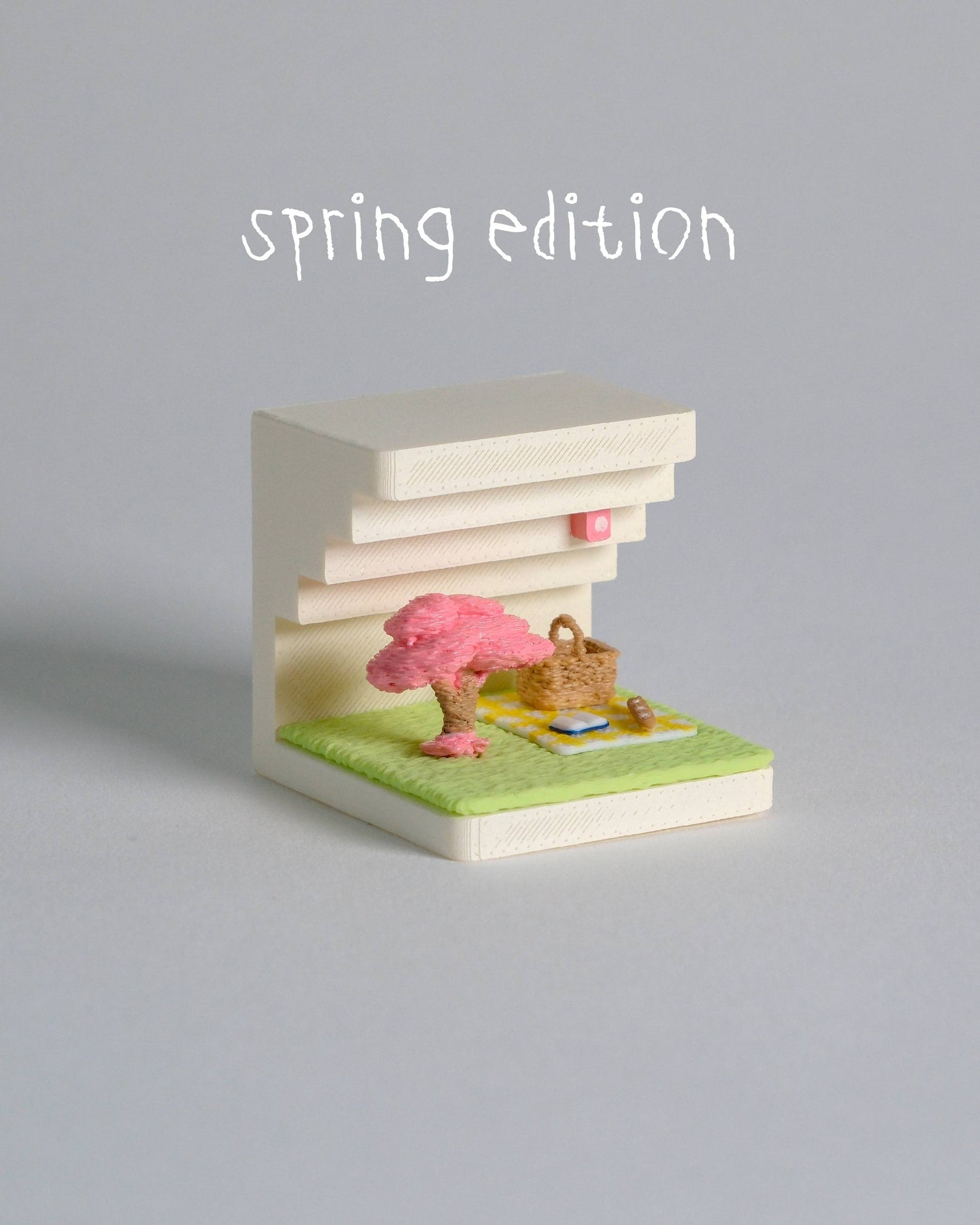 Small model of a building with a cherry blossom tree and picnic setup on a gray background, labeled 'spring edition'.