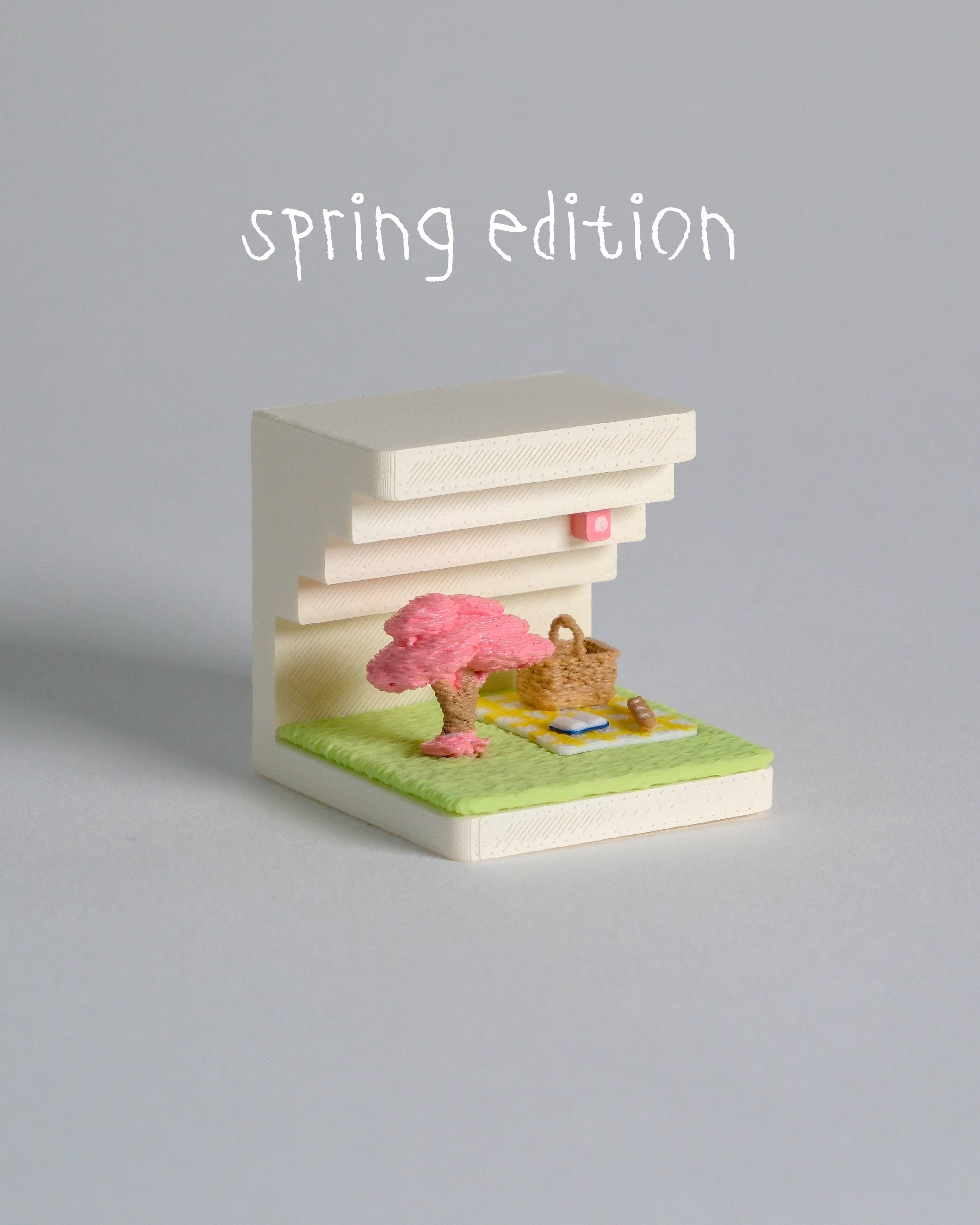 Small model of a building with a cherry blossom tree and picnic setup on a gray background, labeled 'spring edition'.