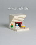 A miniature winter-themed miniature house set with a cream-colored fireplace, a small snowman figure, a yellow-and-white stool, and a red brick fireplace opening, with the text 'winter edition' above on a plain gray background.