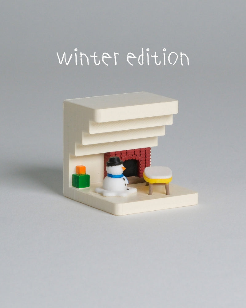 A miniature winter-themed miniature house set with a cream-colored fireplace, a small snowman figure, a yellow-and-white stool, and a red brick fireplace opening, with the text 'winter edition' above on a plain gray background.