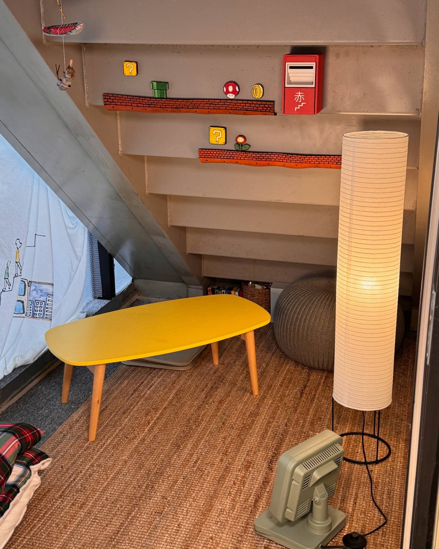 A cozy, compactly furnished room corner featuring a bright yellow oval coffee table, a tall white ribbed floor lamp, a woven brown pouf, and a retro-style green heater on a textured brown rug. In the background, a gray staircase ascends against a beige wall decorated with miniature Mario-themed shelf props.