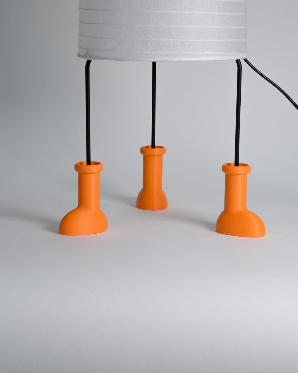 A modern table lamp with a white ribbed drum shade supported by three black metal legs that terminate in bright orange bottle-shaped bases, shown on a light gray studio background.