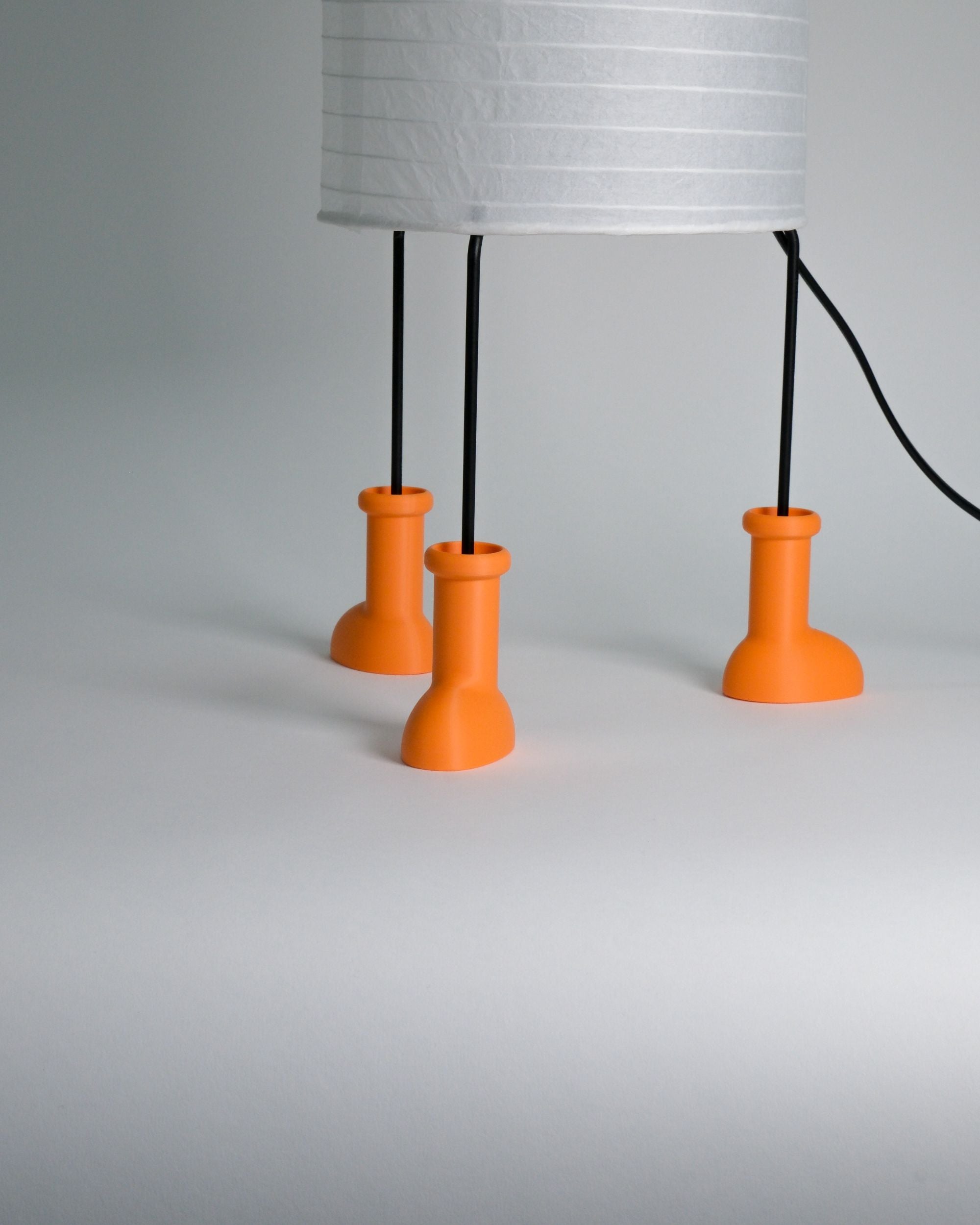 A modern table lamp with a white pleated drum shade supported by three bright orange cylindrical feet, with thin black legs and a black power cord, shown on a neutral gray background.